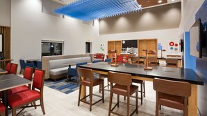 A modern dining area with a long table, red and brown chairs, a blue ceiling feature, and a kitchen area in the background. - Holiday Inn Express & Suites Minneapolis – West End by IHG (Golden Valley)