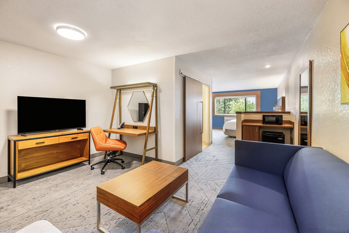 A modern living room with a flat-screen TV, a wooden desk, an orange office chair, a blue sofa, and a kitchen area with a microwave.