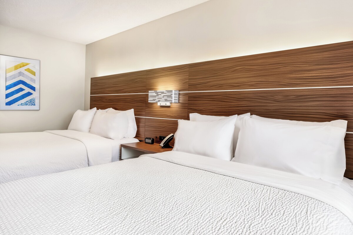 A hotel room with two beds, a wooden headboard, a bedside table with a lamp, and a wall-mounted light fixture.