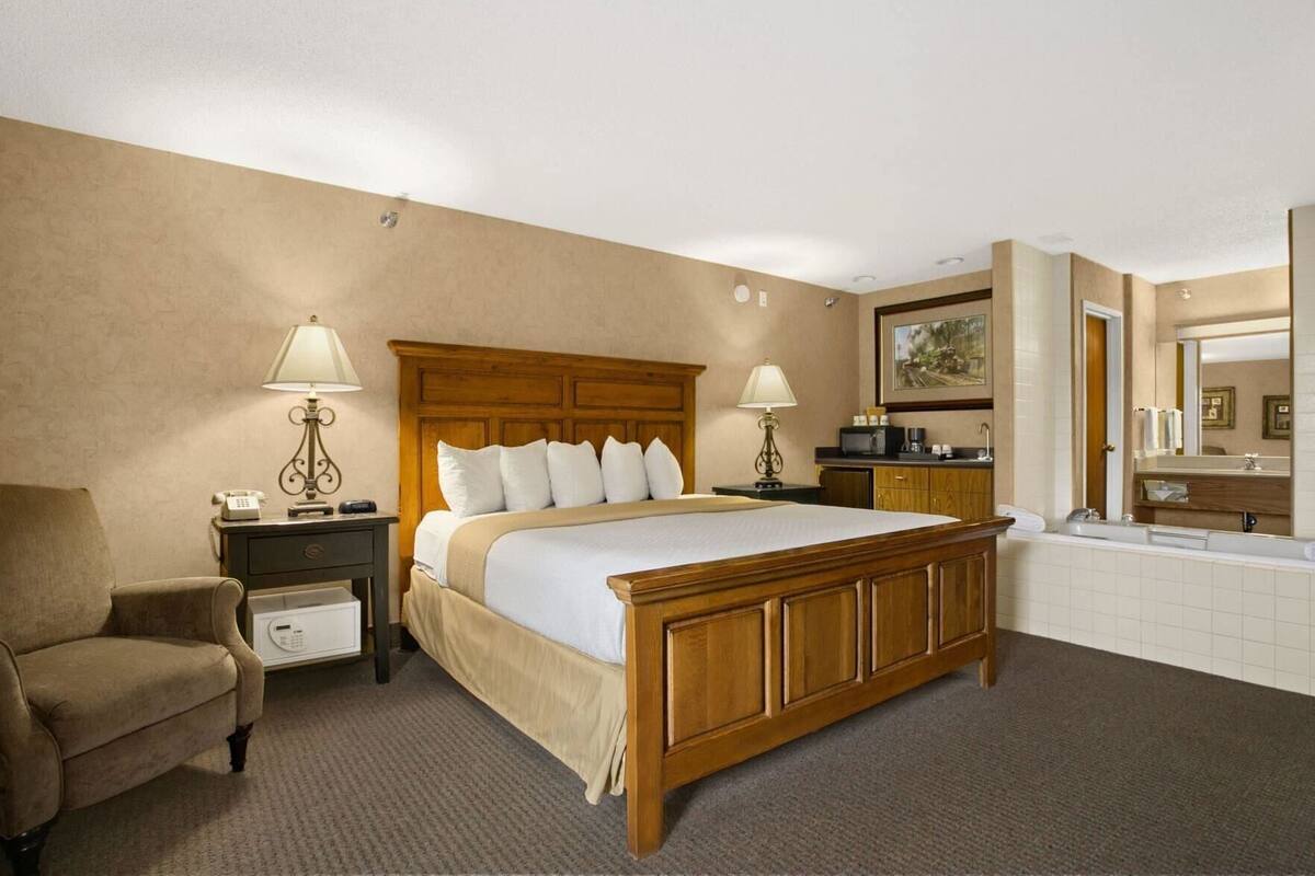 Une chambre d’hôtel avec un lit en bois, deux tables de chevet avec des lampes, une chaise, un petit bureau, une salle de bain avec un lavabo et un miroir, et une grande fenêtre.