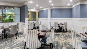 A hotel dining area with multiple tables and chairs, large windows with plants, and a patterned carpet.