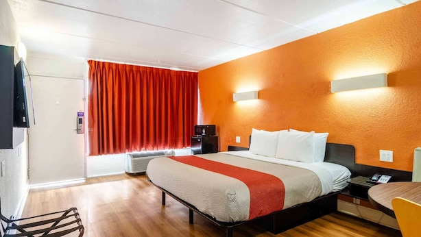 A hotel room with a bed, a desk, a chair, an air conditioning unit, and a window with red curtains. - Motel 6 Brunswick, GA (Brunswick)