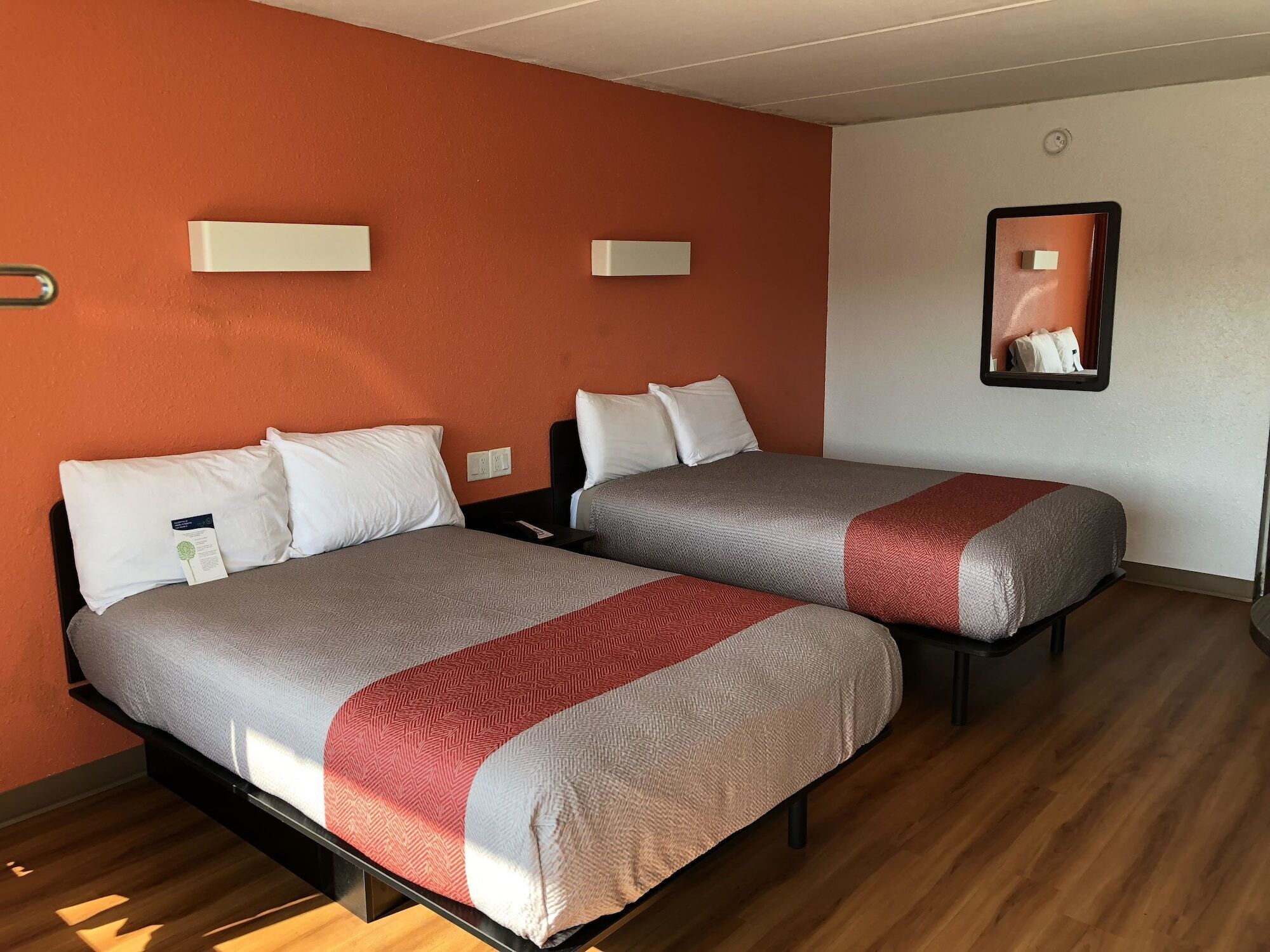 A hotel room with two beds, each with a grey and red blanket, white pillows, and a small bedside table with a lamp.