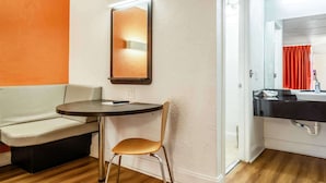 A hotel room with a white sofa, a round table, a chair, a mirror, and a bathroom with a sink and red curtains. - Motel 6 Brunswick, GA (Brunswick)
