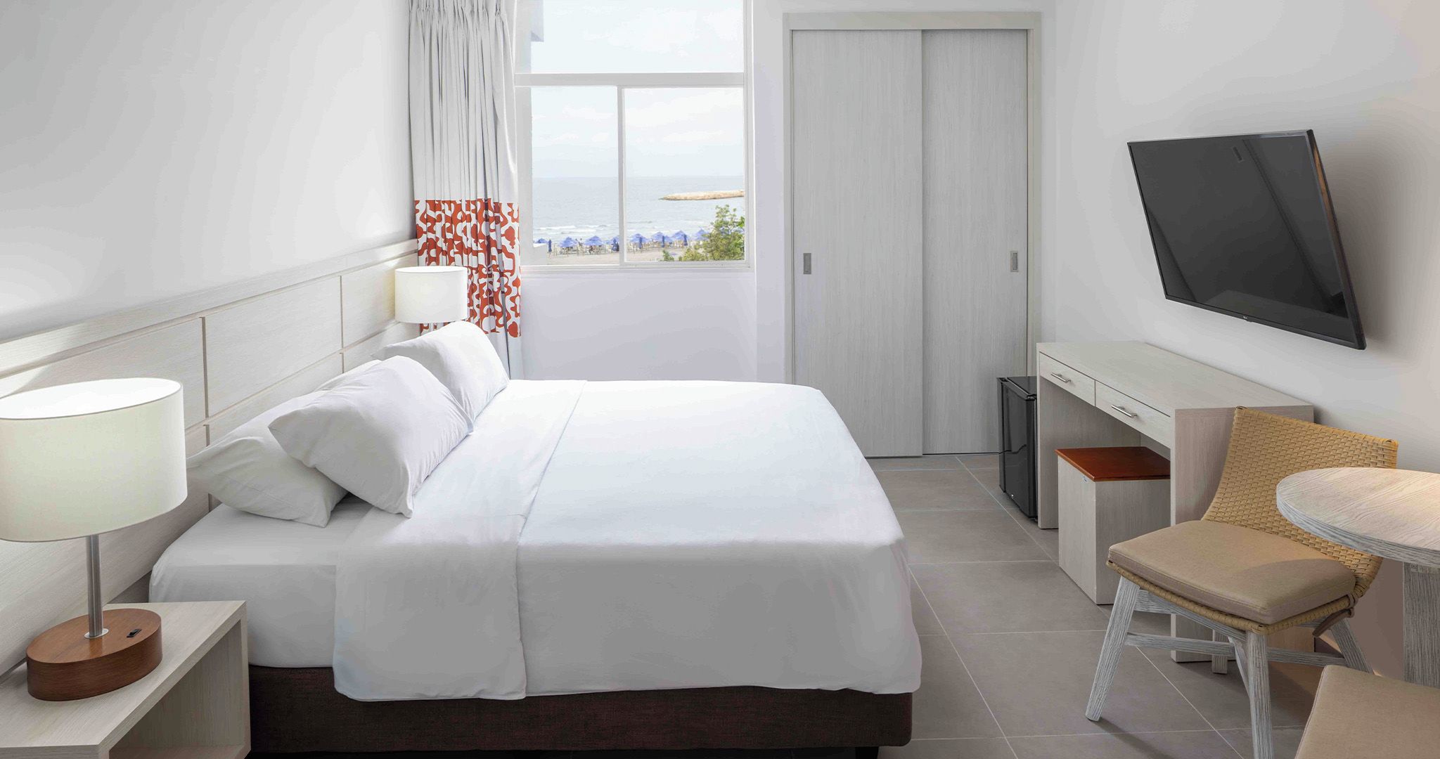 Habitación de hotel con cama, mesita de noche, lámpara, televisión y vistas a la playa.