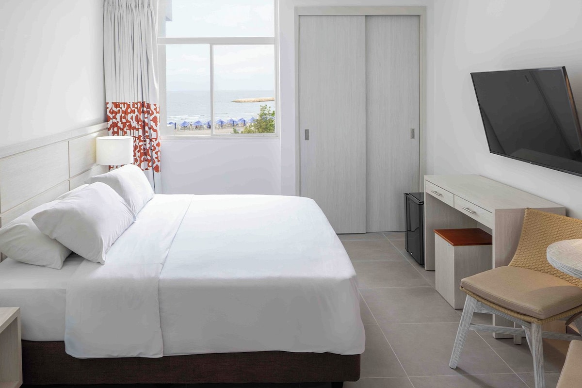 Ein Hotelzimmer mit Bett, Nachttisch, Lampe, Fernseher und Blick auf den Strand.
