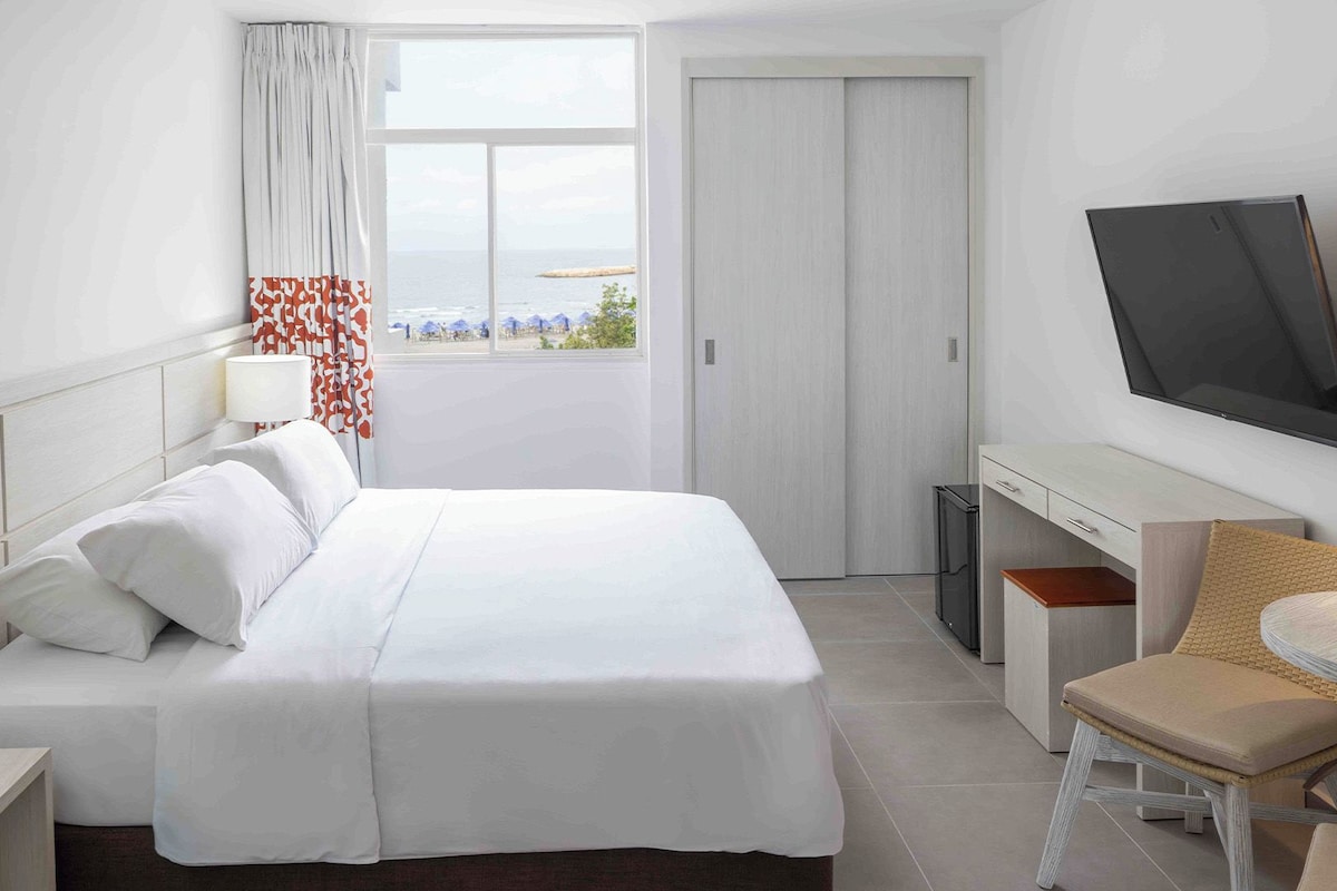 Ein Hotelzimmer mit Bett, Schreibtisch, Stuhl, Fernseher und Fenster mit Meerblick.
