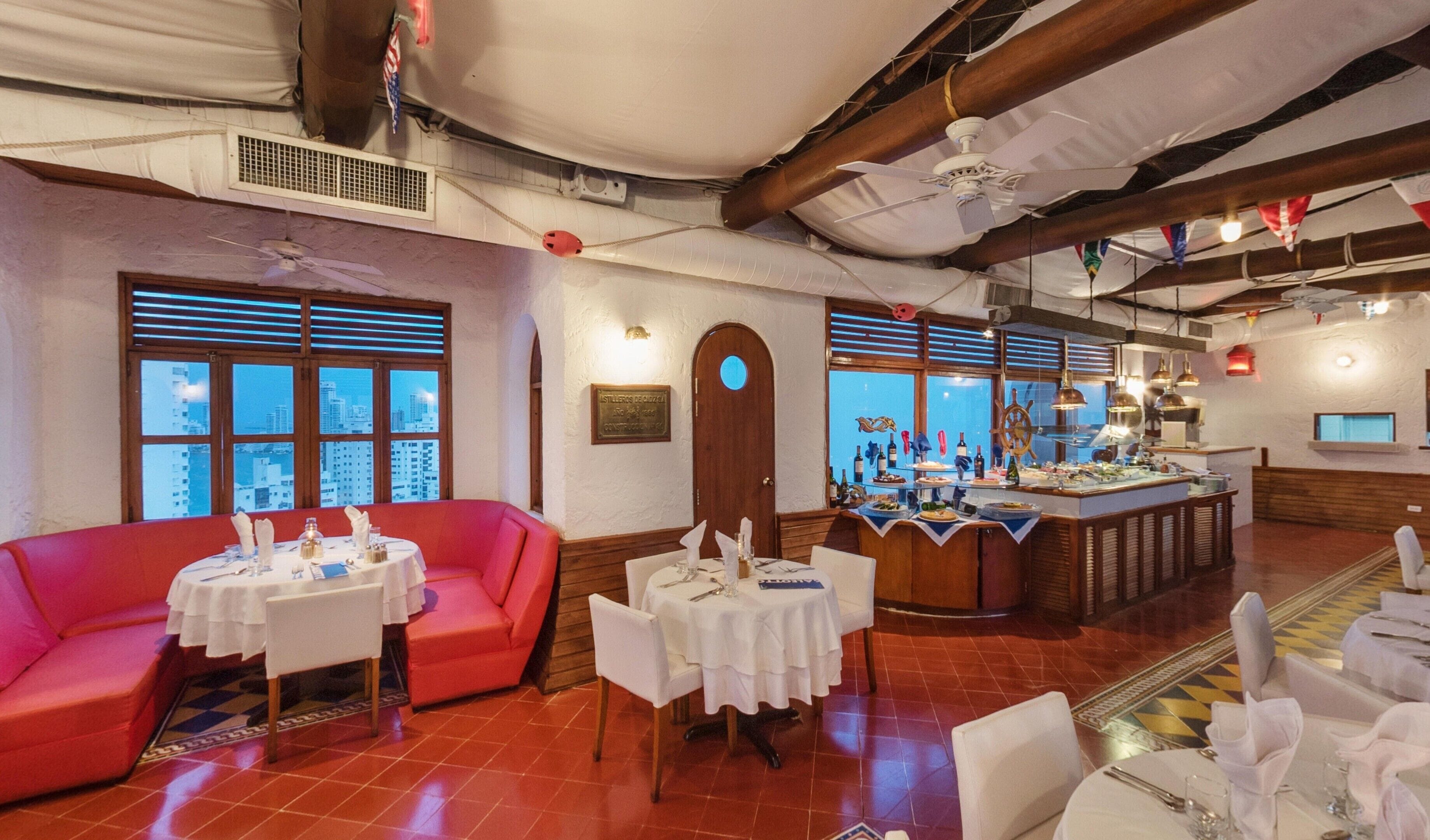 Un restaurante con asientos rojos, mesas blancas y una zona de bar con botellas y una rueda de barco.