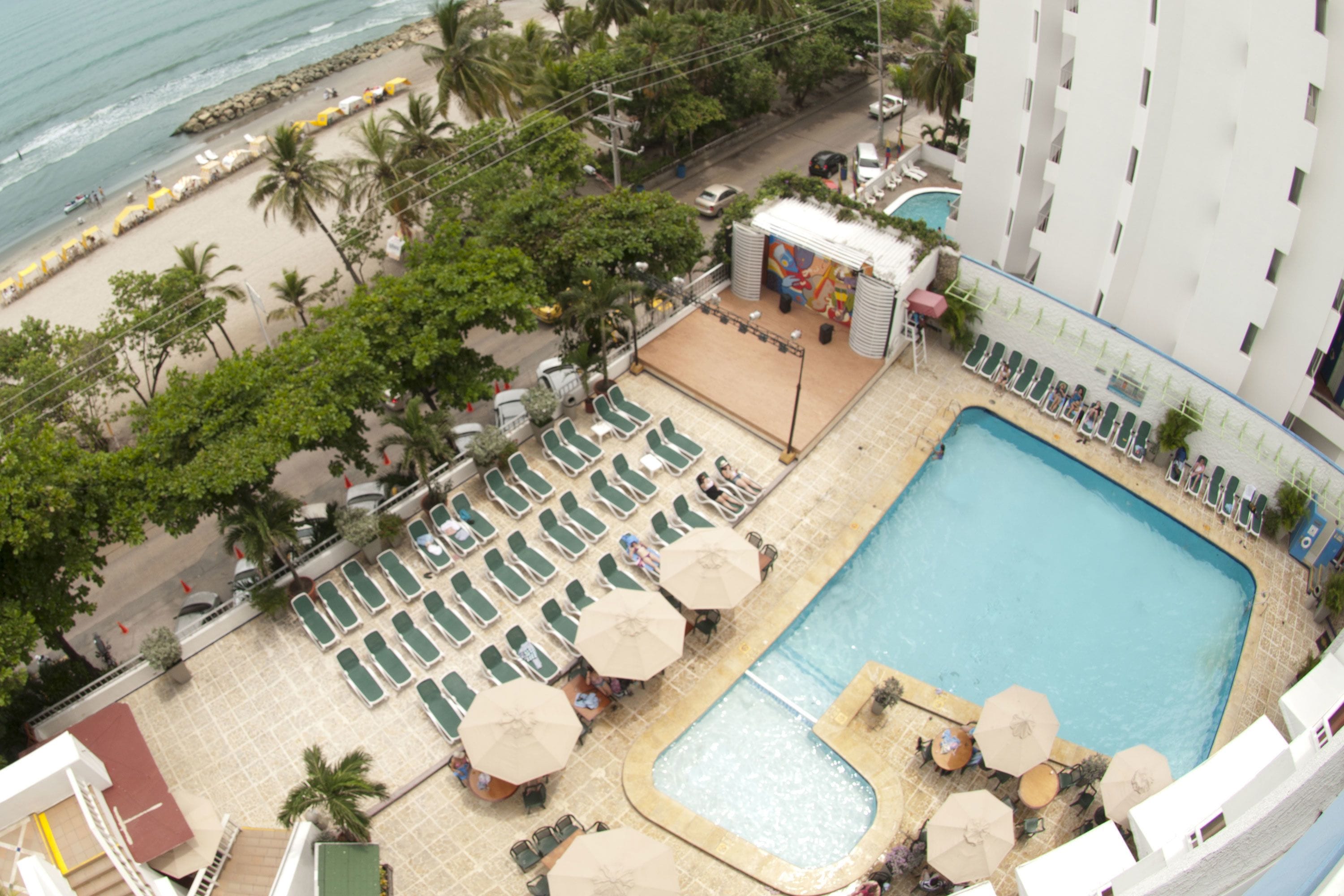 An aerial view of a hotel with a swimming pool, sun loungers, umbrellas, and a beach area with umbrellas and lounge chairs.