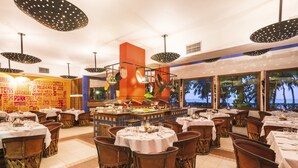 Ein Restaurant mit runden Tischen, Korbstühlen und einer Bar in der Mitte, mit großen Fenstern und Blick auf Palmen.