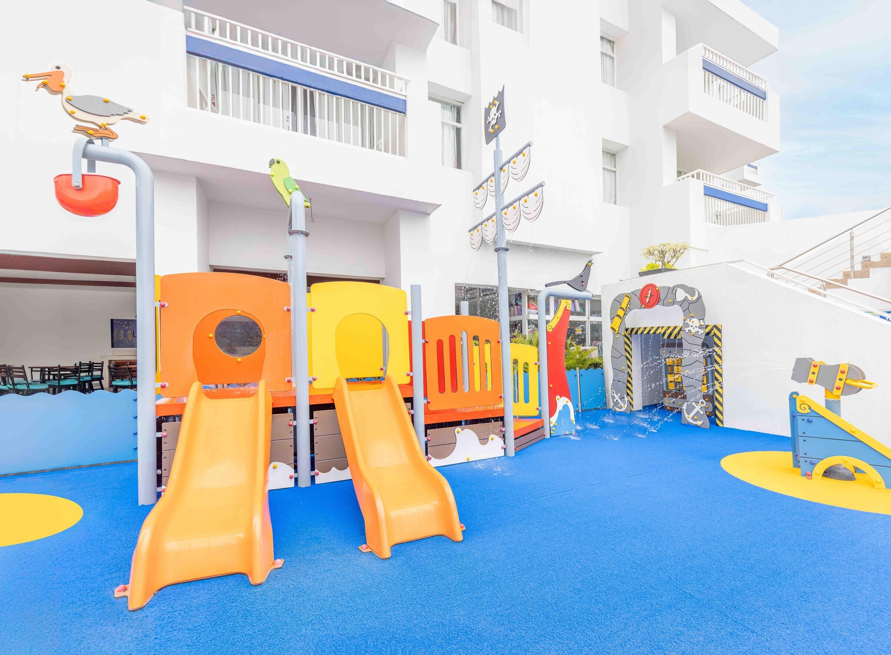 A children's play area with slides and climbing structures, adjacent to a white building with balconies.