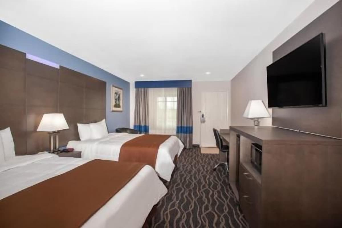 A hotel room with two beds, a desk, a flat-screen TV, and a large window with curtains.