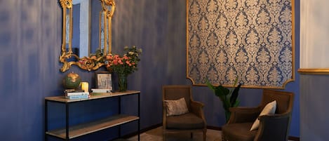 A room with a large ornate mirror, a console table with books and a globe, two upholstered chairs, and a patterned wallpaper.