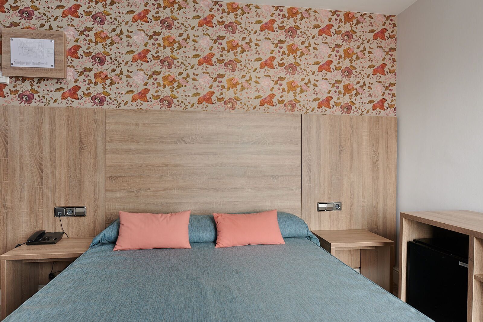 A bedroom with a wooden headboard, two bedside tables, a bed with a blue bedspread and two pink pillows, and a floral wallpaper.