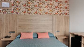 A bedroom with a wooden headboard, two bedside tables, a bed with a blue bedspread and two pink pillows, and a floral wallpaper.