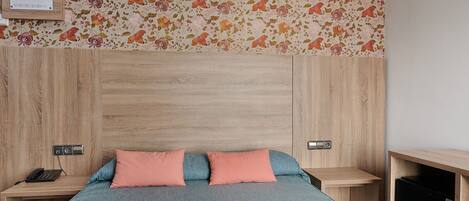 A bedroom with a wooden headboard, two bedside tables, a bed with a blue bedspread and two pink pillows, and a floral wallpaper.