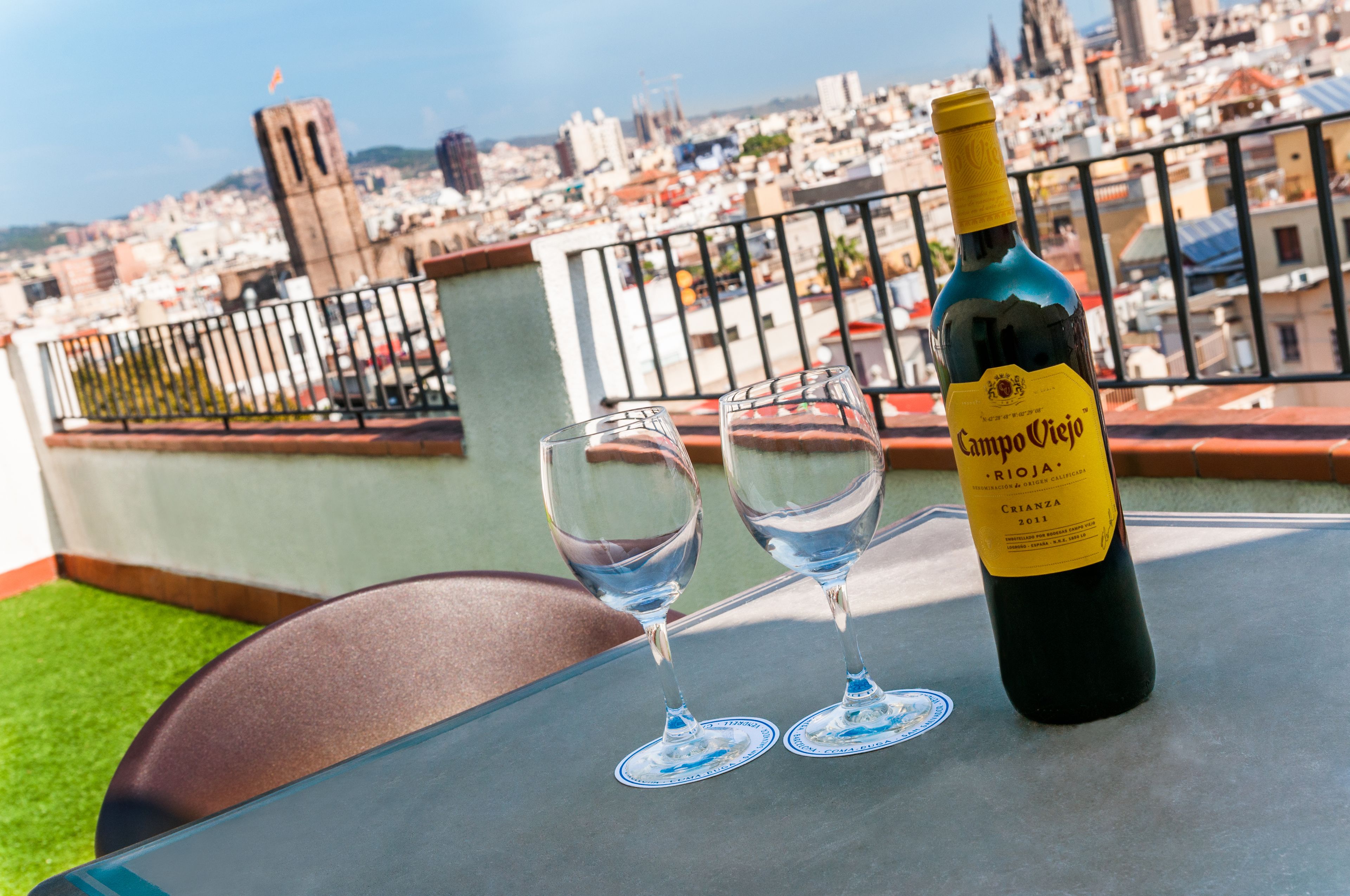 Una bottiglia di vino Campo Viejo Rioja Crianza 2011 su un tavolo, insieme a due bicchieri da vino vuoti, con vista su un panorama urbano.