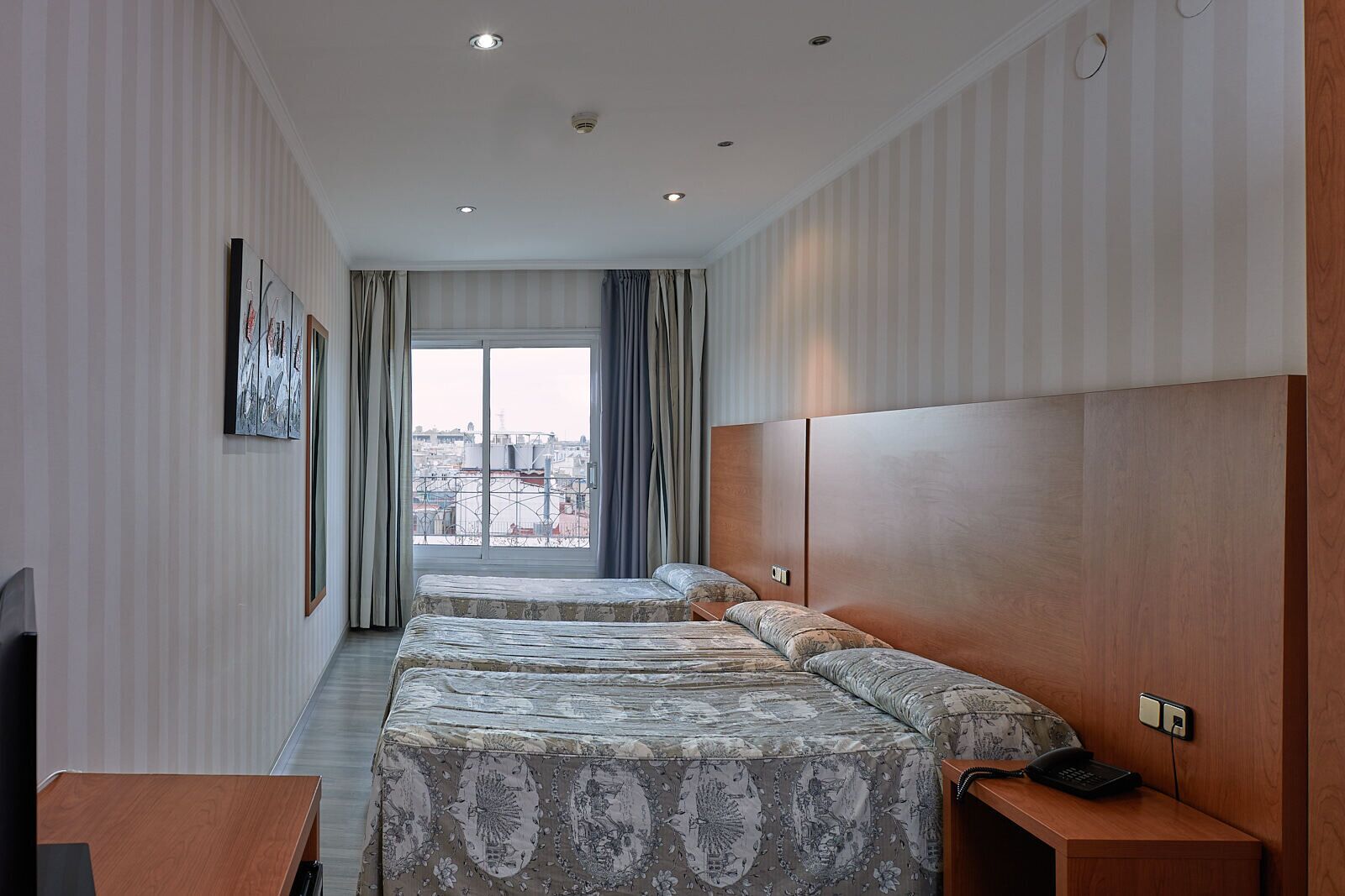 A hotel room with two beds, a wooden headboard, a desk, and a view of the city through large windows.
