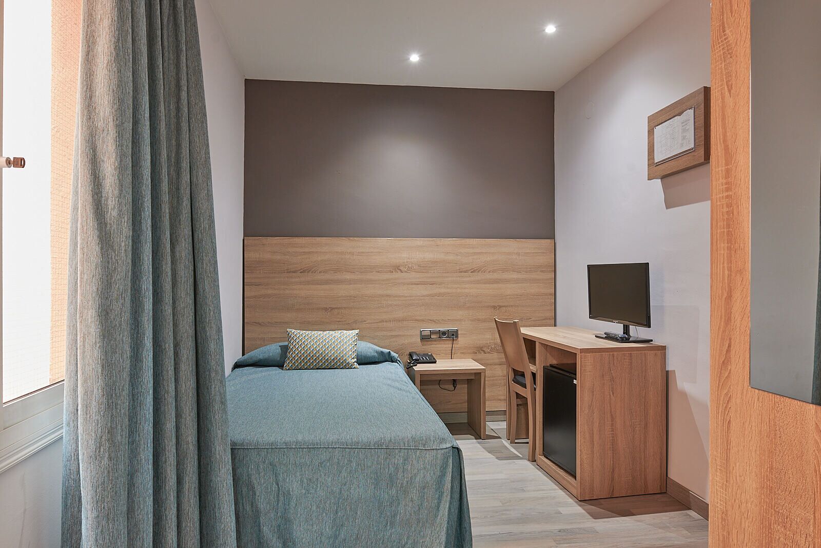 A small, modern hotel room with a bed, a desk, a chair, a TV, and a wooden headboard.