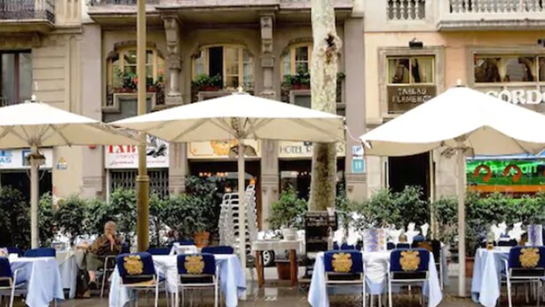 Un café al aire libre con mesas y sombrillas, frente a un edificio con letreros visibles.