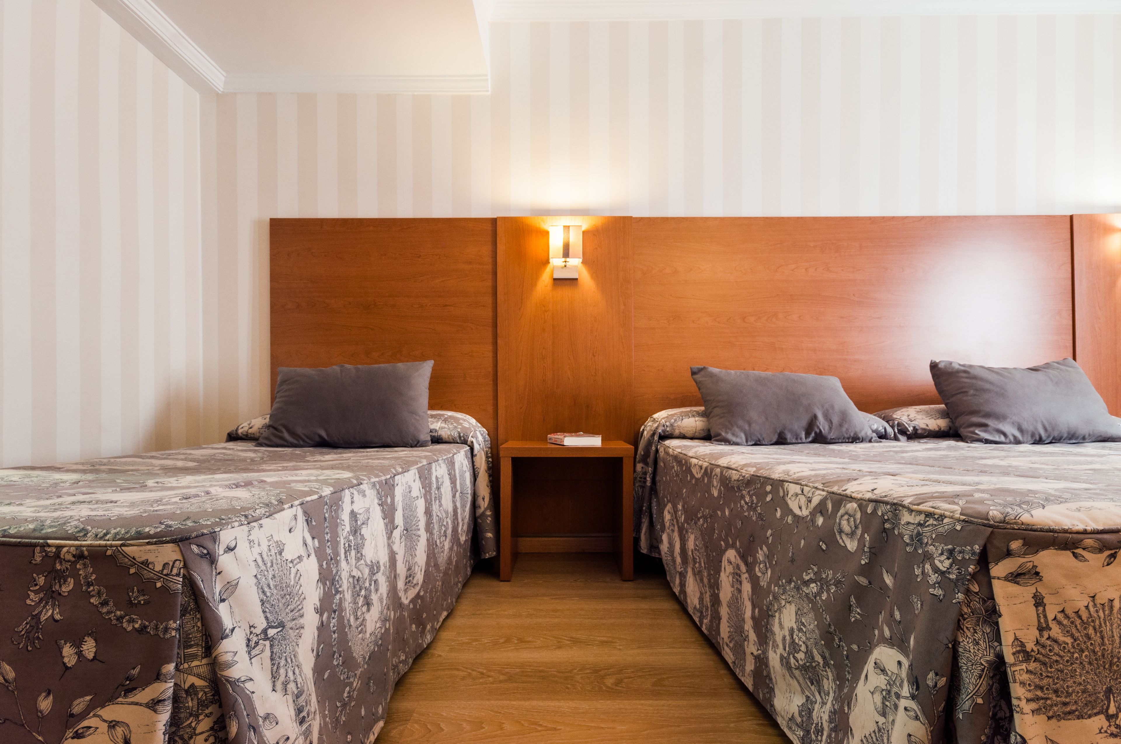 A hotel room with two single beds, a wooden headboard, a nightstand, and a wall-mounted light.