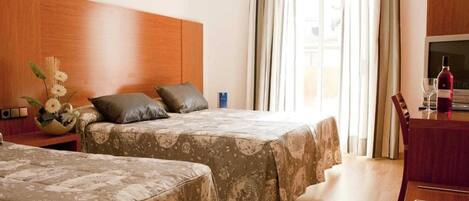 A hotel room with two beds, a wooden headboard, a desk with a bottle and glasses, and a window with curtains.