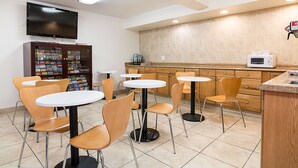 Un área de cocina limpia y moderna, con microondas, cafetera y refrigerador. Varias mesas redondas y sillas dispuestas para comer.