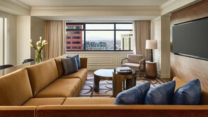 Habitación de hotel moderna con un sofá grande, una mesa redonda de centro y un televisor de pantalla plana. Se aprecia una vista de la ciudad a través de amplias ventanas.