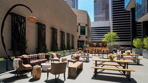 Un espacio al aire libre con sillón de cuero, sillas y mesas de madera, rodeado de edificios altos.