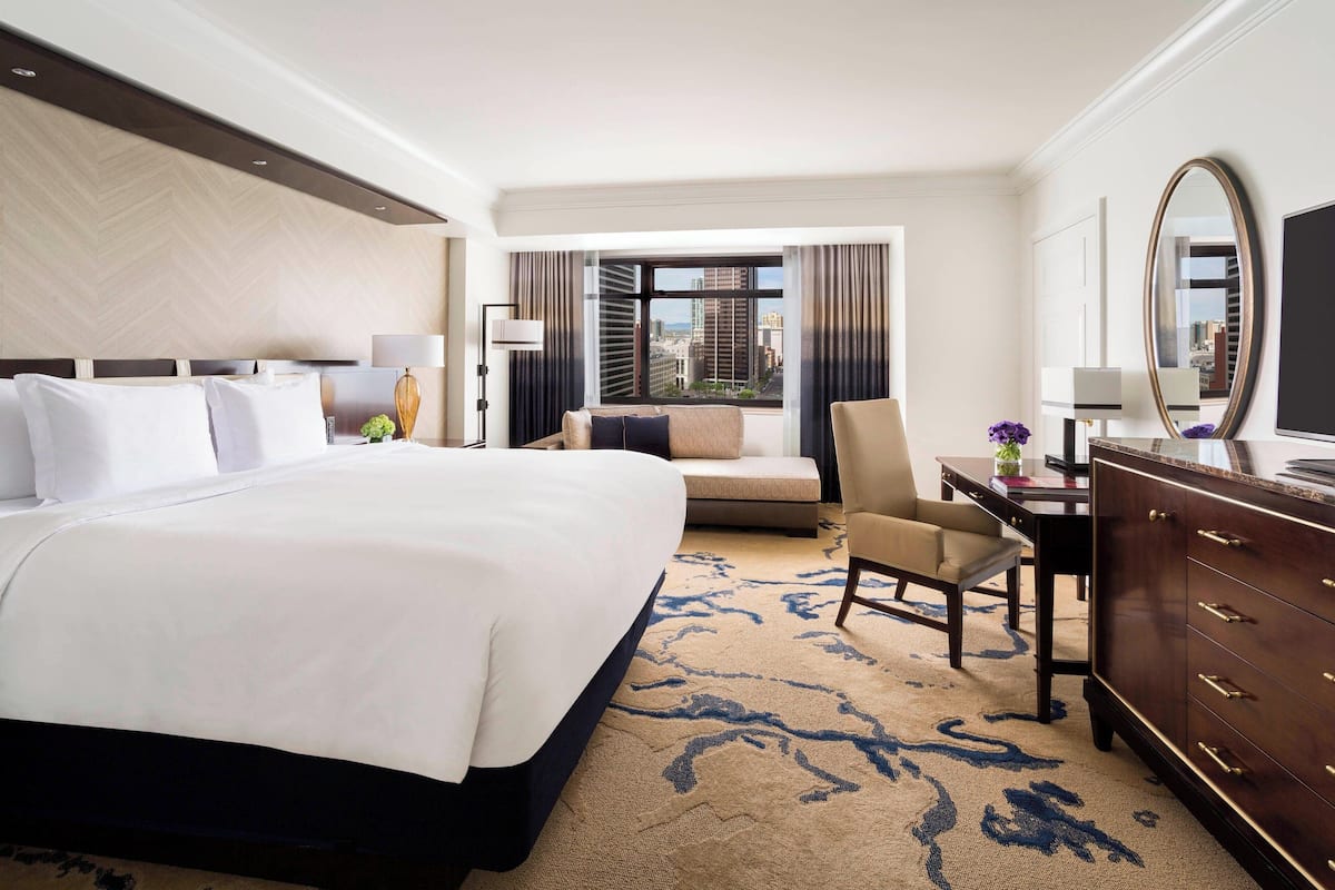 A hotel room with a large bed, a desk, a chair, and a dresser. There is a window with a view of buildings outside.