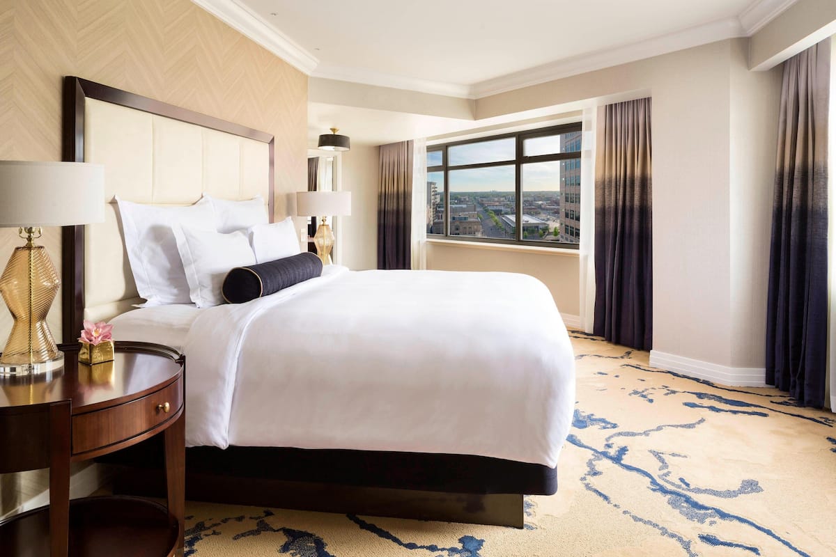 A hotel room with a large bed, a nightstand, a lamp, and a view of the city through a window.