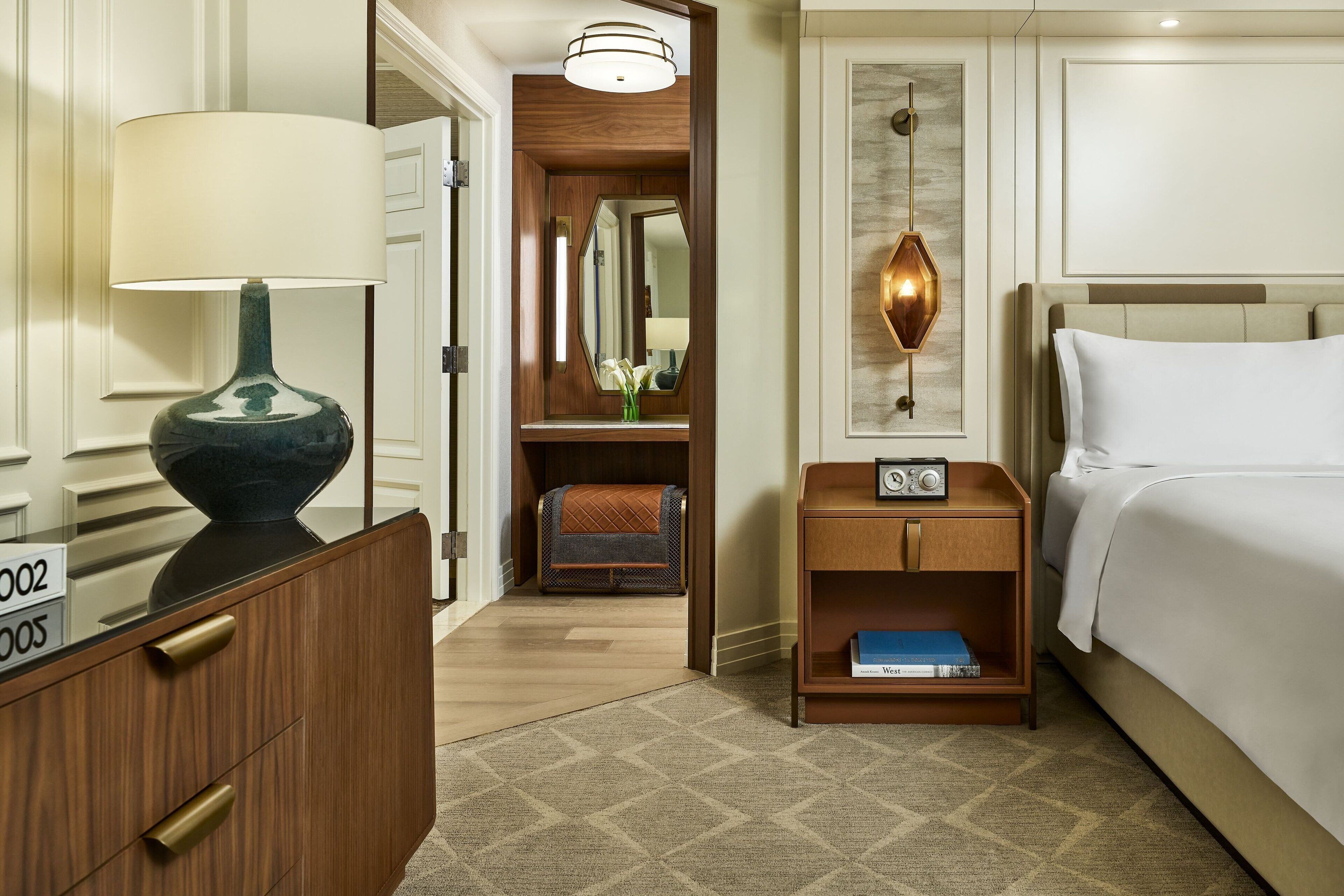 A hotel room with a bed, bedside table, lamp, and a small shelf with books.