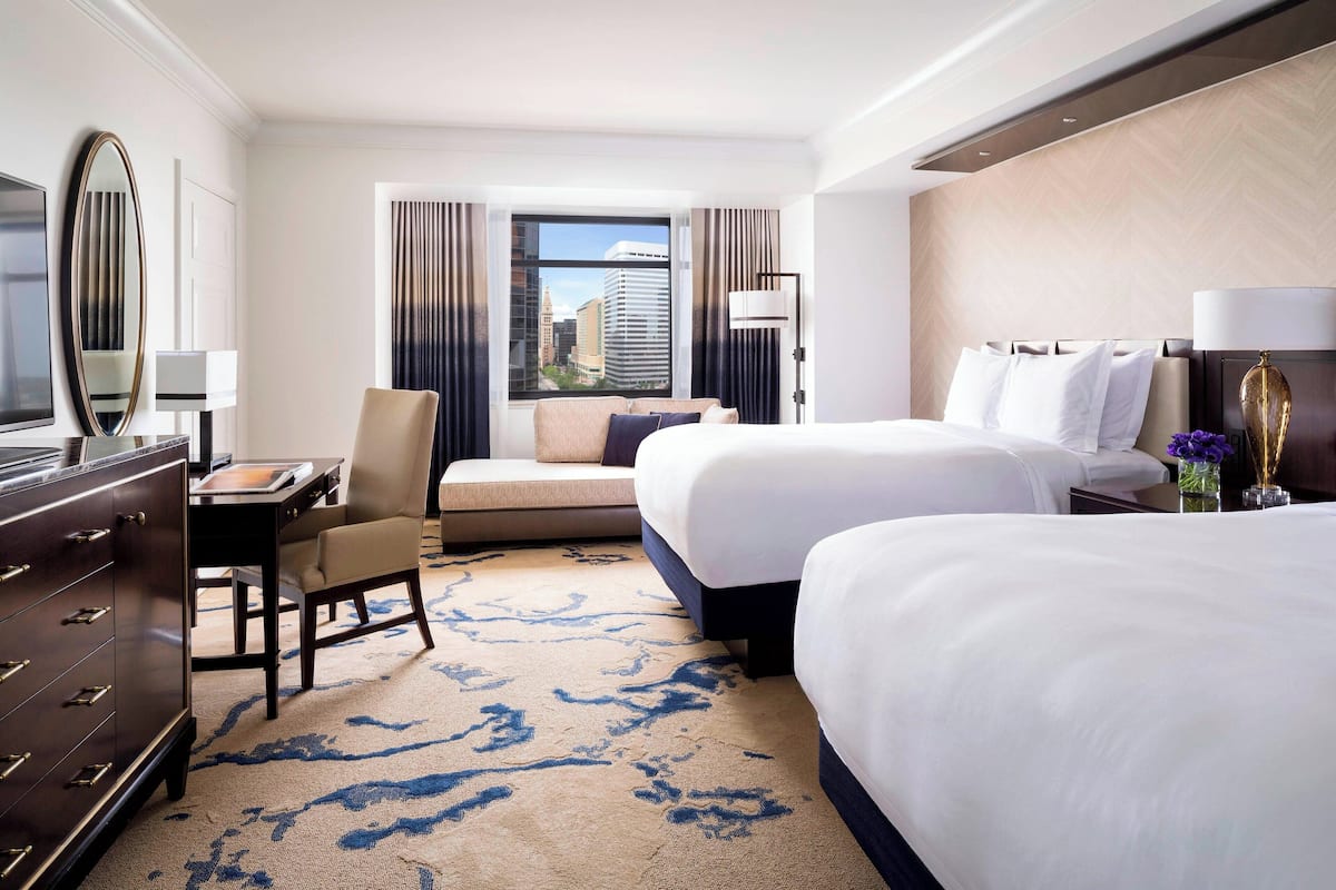 A hotel room with two beds, a desk, a chair, a dresser, and a large window with a city view.