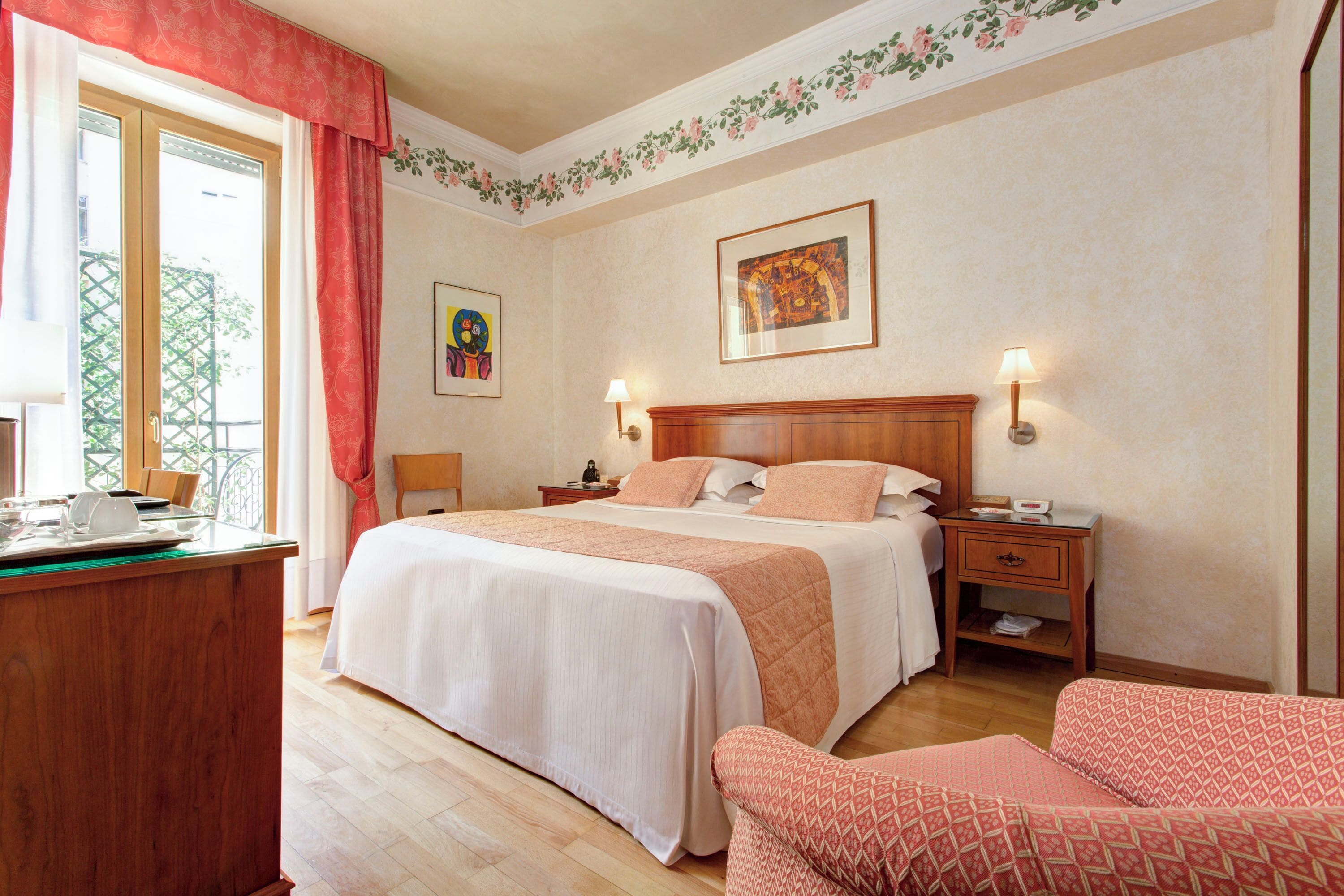 A hotel room with a large bed, a desk with a chair, a nightstand, and a red patterned armchair.