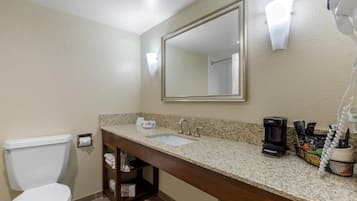 Une salle de bain avec des toilettes blanches, un lavabo avec un plan de travail, un miroir, une machine à café et un sèche-cheveux.