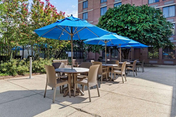 Outdoor patio seating with round tables and chairs, blue umbrellas, and a brick building in the background.