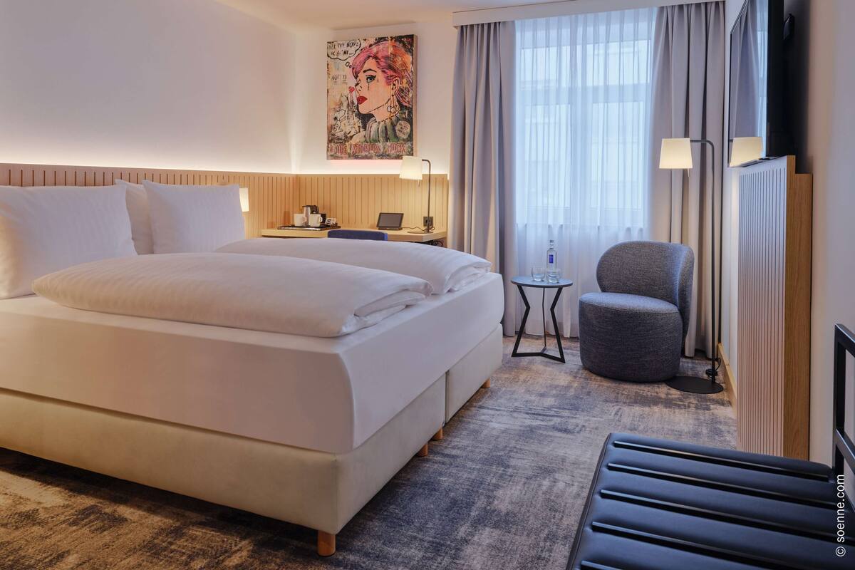 A hotel room with a large bed, a small table with a laptop, a chair, and a painting on the wall.