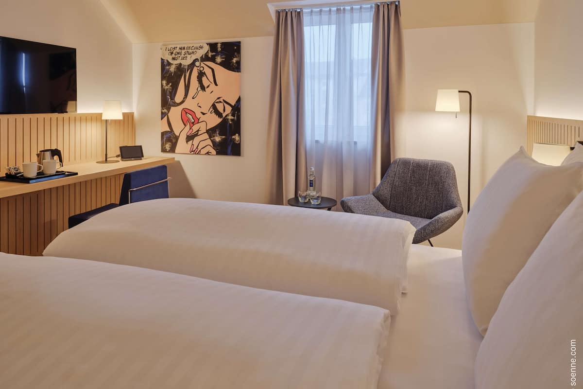 A hotel room with two beds, a desk with a laptop, a chair, a TV, and a painting on the wall.