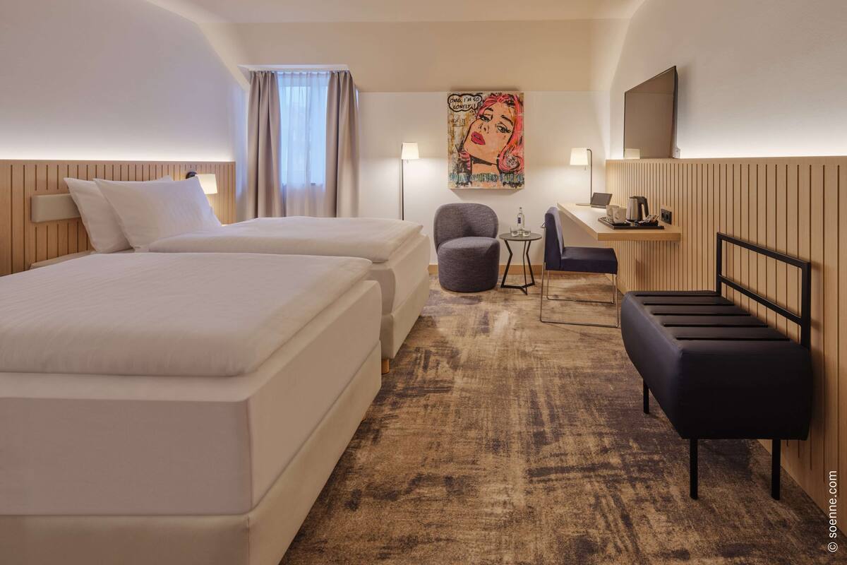 A hotel room with two beds, a desk, a chair, a small table, and a painting on the wall.