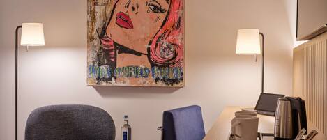Habitación de hotel con una obra de arte grande en la pared, una mesa redonda con una cafetera, dos sillas y una botella de agua sobre una mesita.