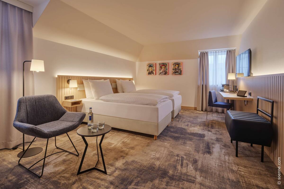 A hotel room with a bed, a desk, a chair, a small table with glasses and a bottle, and a lamp.