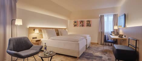 A hotel room with a bed, a desk, a chair, a small table with glasses and a bottle, and a lamp.