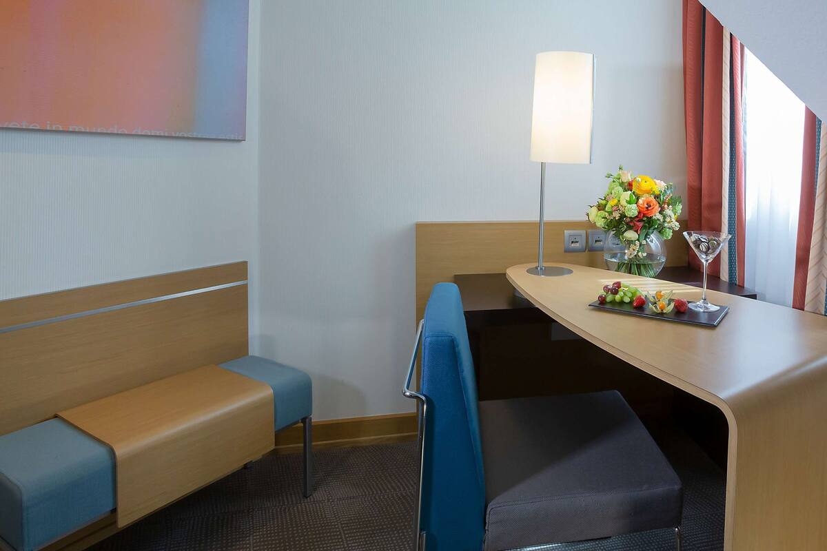 A hotel room with a bed, a desk with a lamp, a vase of flowers, and a fruit tray.