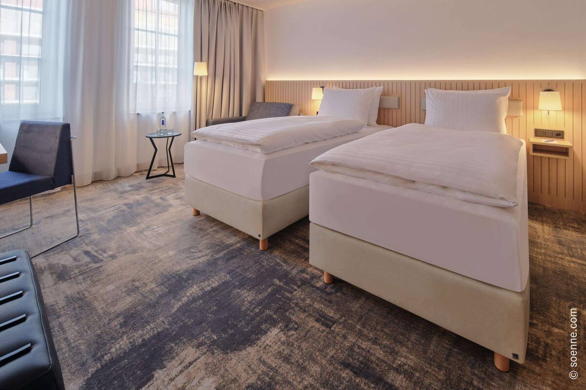 A hotel room with two beds, a desk, a chair, and a view of the outside through large windows.
