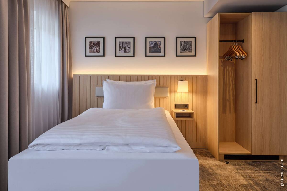 A hotel room with a neatly made bed, a bedside table with a lamp, a wardrobe, and four framed pictures on the wall.