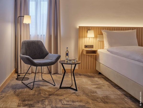 Una habitación de hotel con una cama, una silla, una mesita pequeña y una mesita de noche con lámparas.