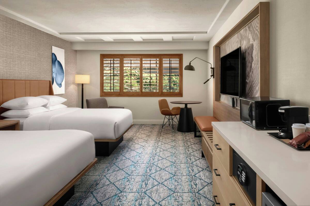 A hotel room with two beds, a desk with a TV, a coffee maker, and a window with wooden shutters.