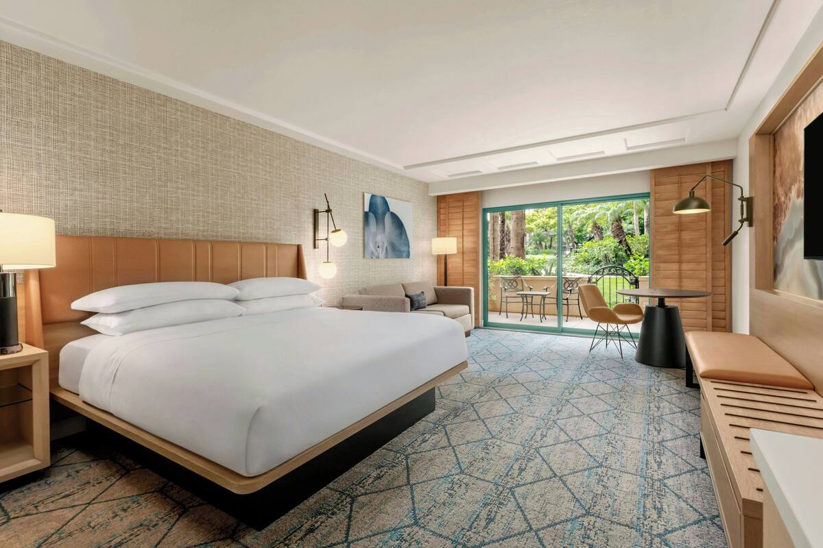 A hotel room with a large bed, a desk with chairs, a TV, and a view of greenery through the window.