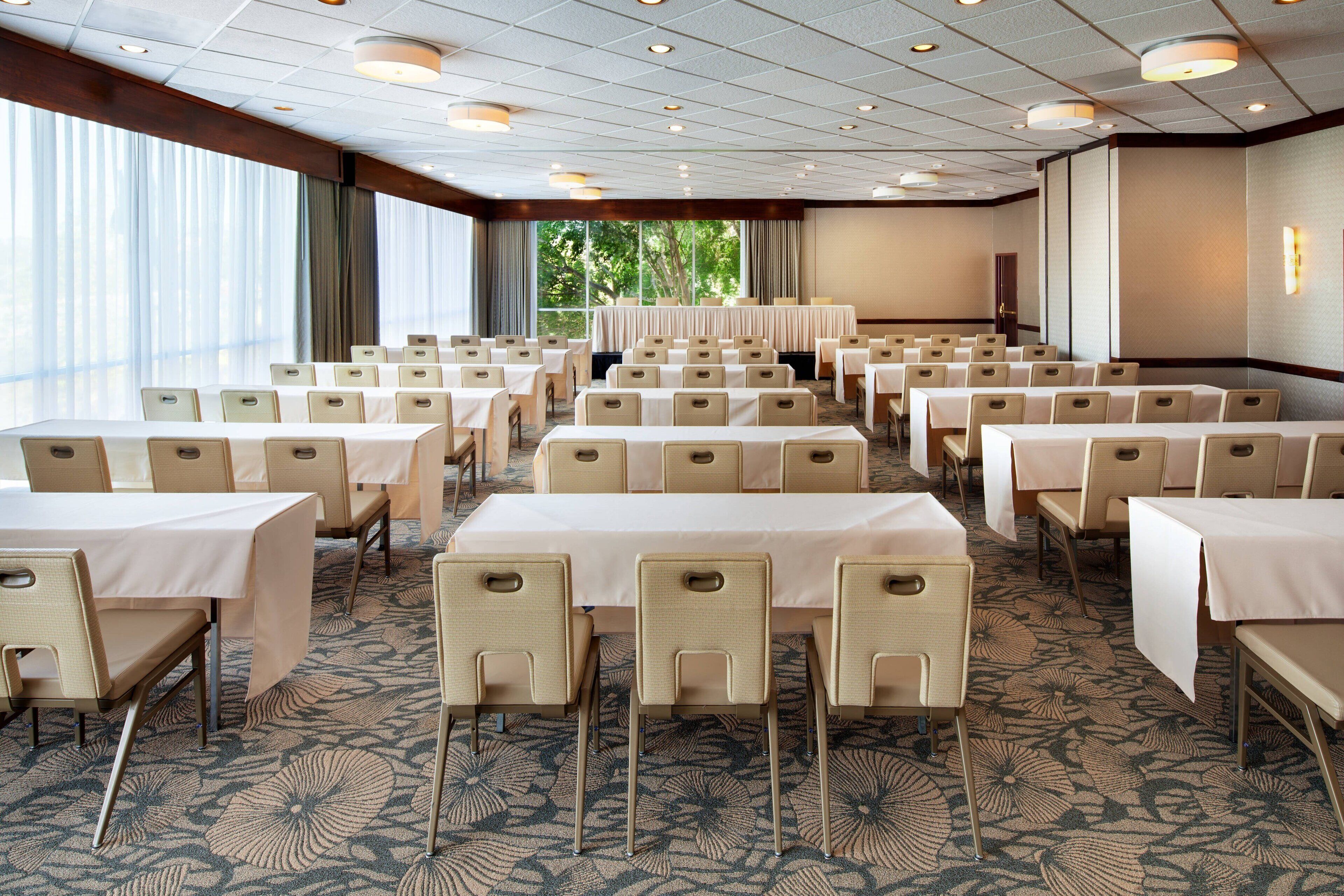 A conference room with rows of tables and chairs, large windows, and a carpeted floor.