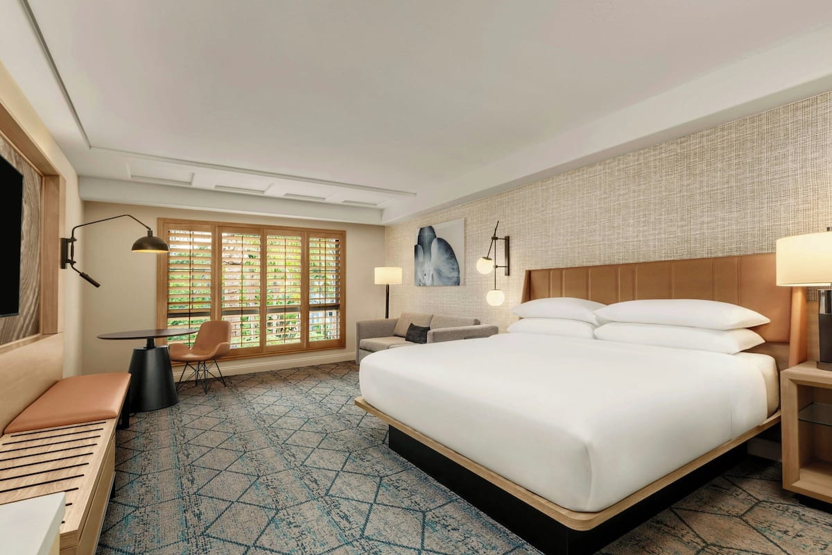 A hotel room with a large bed, a desk, a chair, and a window with blinds.